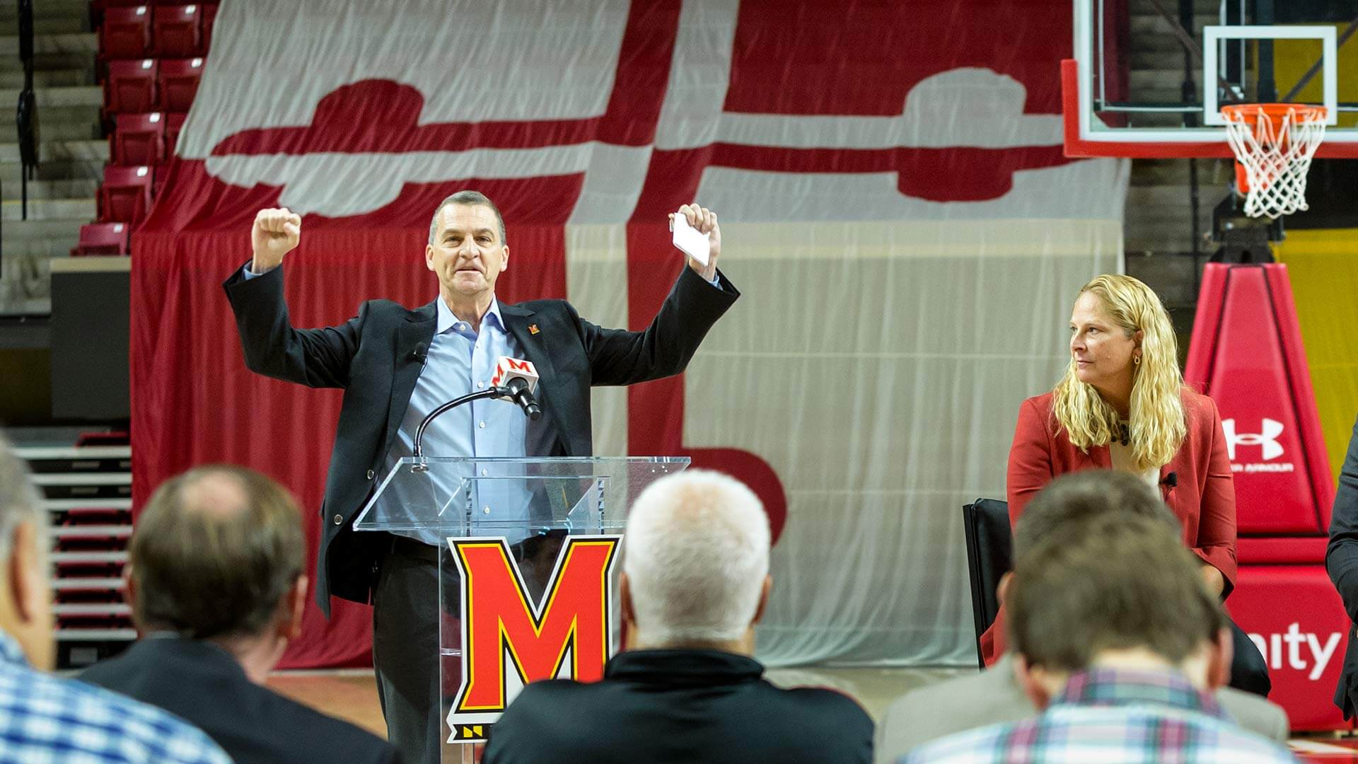 Men's head basketball coach Mark Turgeon raises his arms at the podium next to women's head coach Brenda Freese